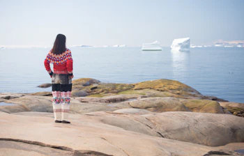 Ung inuitkvinde i farverig traditionel grønlandsk dragt står på klippekysten ved Ilulissats isfjord med isbjerge i baggrunden