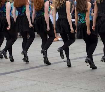 Gruppe af kvindelige irske dansere i farverige traditionelle kostumer viser synkroniserede benbevægelser under en udendørs optræden