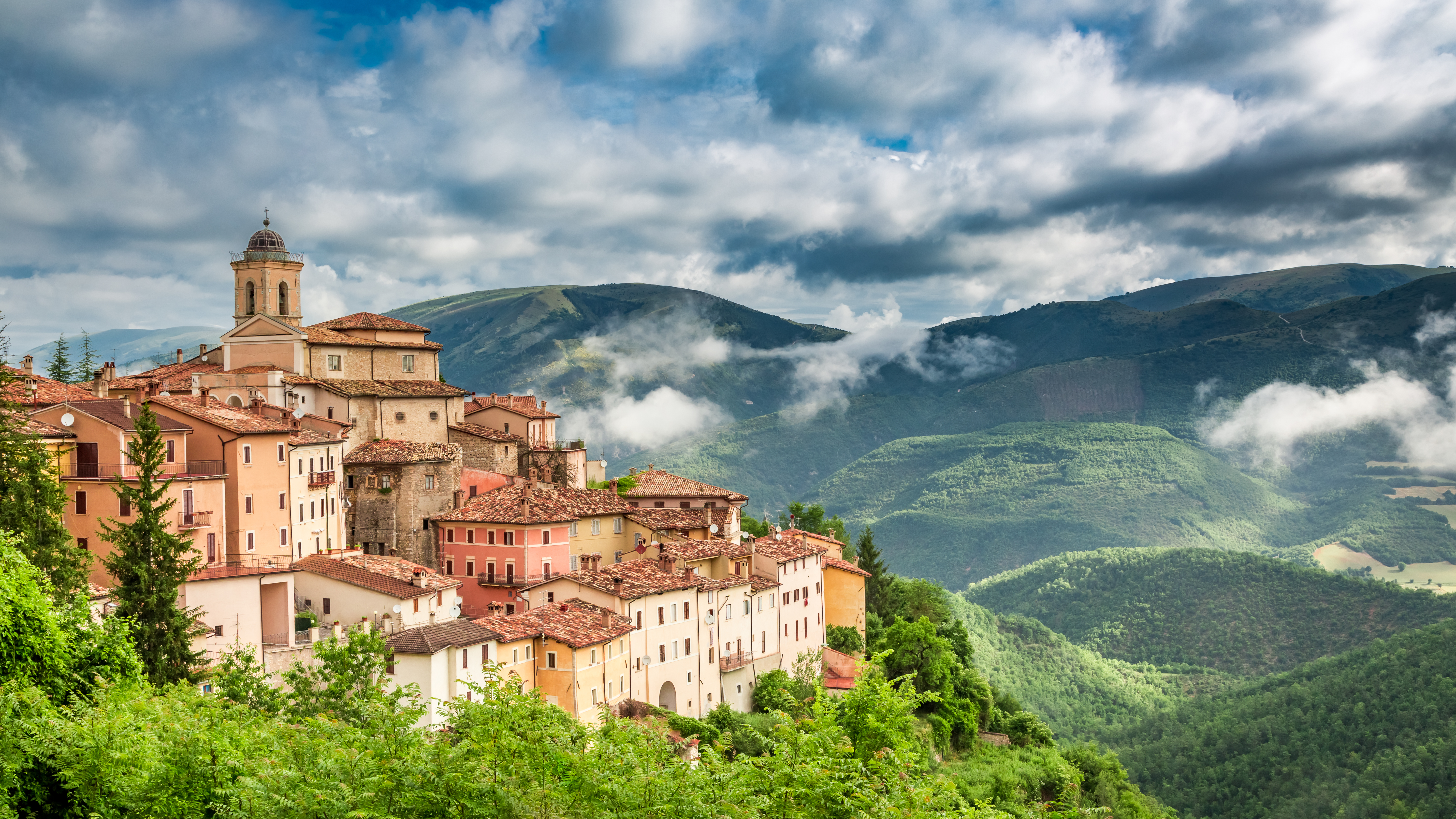 Malerisk italiensk bjerby i Umbria med røde tegltagstage omgivet af grønne bjerge og dramatisk himmel