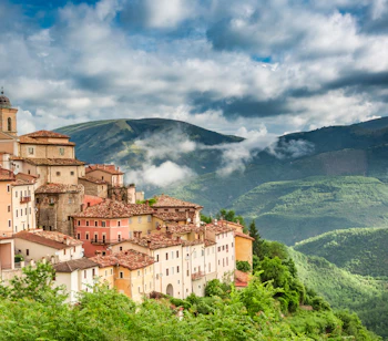 Malerisk italiensk bjerby i Umbria med røde tegltagstage omgivet af grønne bjerge og dramatisk himmel