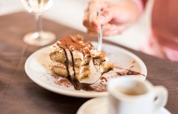 Ung kvinde nyder traditionel italiensk tiramisu-dessert med espressokaffe og et glas hvidvin ved et rustikt træbord på en autentisk italiensk café