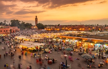 Den berømte Jamaa el Fna markedsplads i Marrakech ved solnedgang med oplyste madboder, folkeliv og Koutoubia-moskeen i baggrunden
