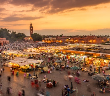 Den berømte Jamaa el Fna markedsplads i Marrakech ved solnedgang med oplyste madboder, folkeliv og Koutoubia-moskeen i baggrunden