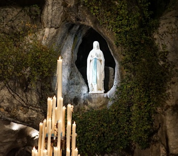 Statue af Jomfru Maria i den hellige grotte i Lourdes, Frankrig, med bedende lys i forgrunden, et berømt katolsk pilgrimssted