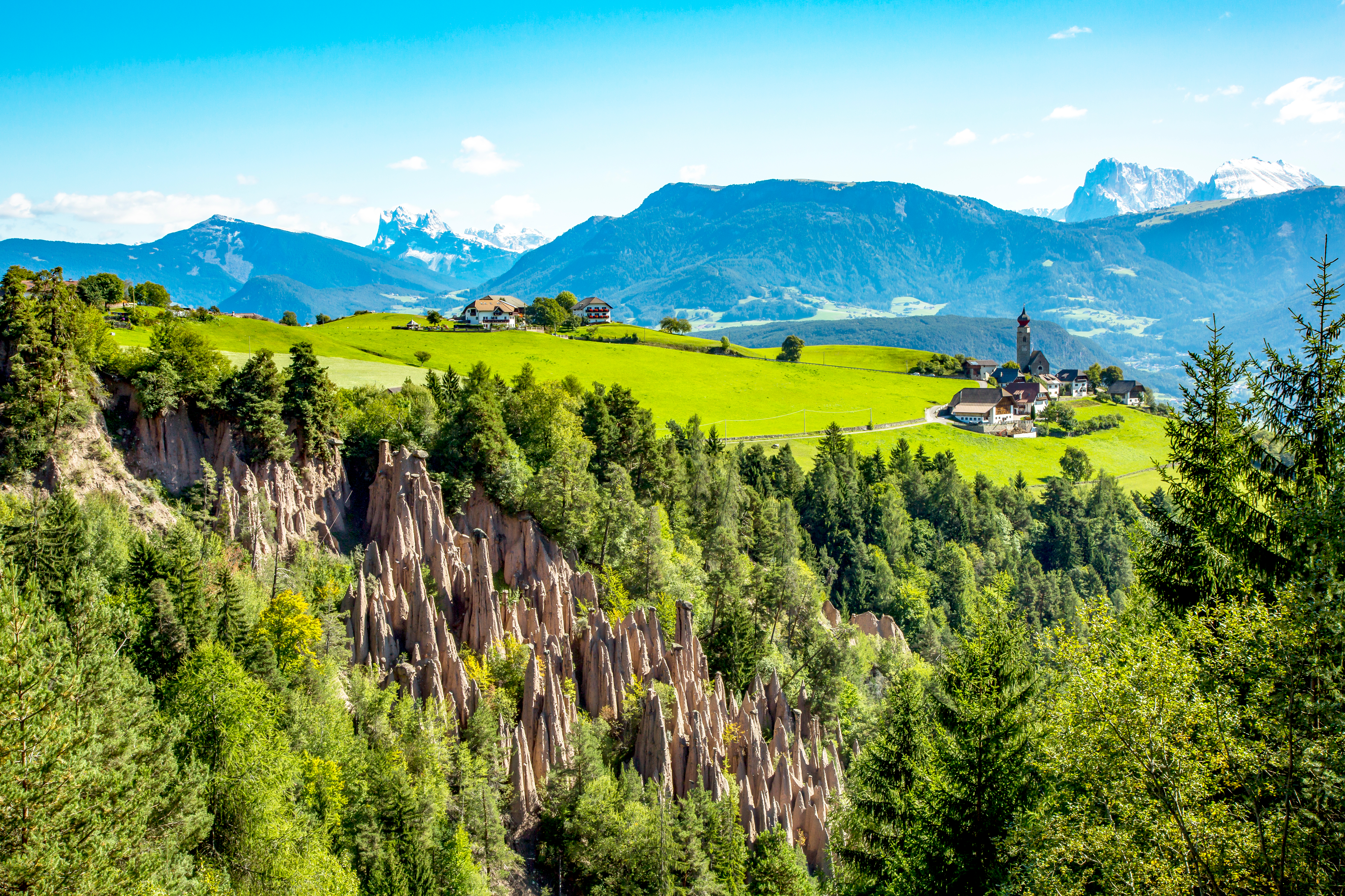Naturlige jordpyramider ved Ritten i Sydtyrol med grønne enge, en lille alpelandsby med kirke og snedækkede Dolomitter i baggrunden