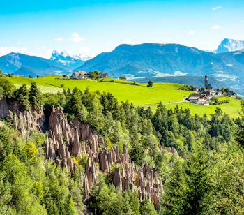 Naturlige jordpyramider ved Ritten i Sydtyrol med grønne enge, en lille alpelandsby med kirke og snedækkede Dolomitter i baggrunden