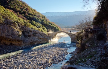 Den historiske Kadiut-stenbro i Benje, hvor lokale krydser over floden, mens en fisker står i det krystalklare vand nær de naturlige termiske bade i Permet