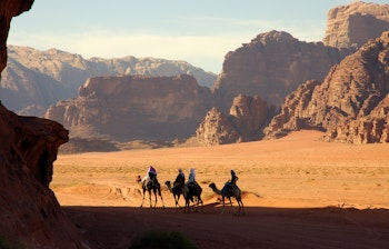 Turister på kamelryggen gennem den spektakulære Wadi Rum ørken i Jordan med røde sandstensklipper og blå himmel