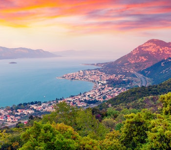 Fugleperspektiv af kystbyen Kamena Vourla i Grækenland ved solnedgang, med farverige bygninger langs Ægæerhavets kyst og bjerge i baggrunden indhyllet i aftendis