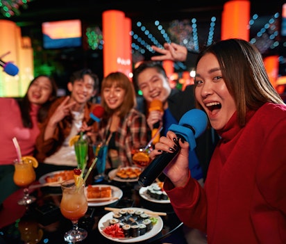 Gruppe af unge venner synger karaoke og tager selfies på en natklub i Seoul med farverige drinks og festlig stemning