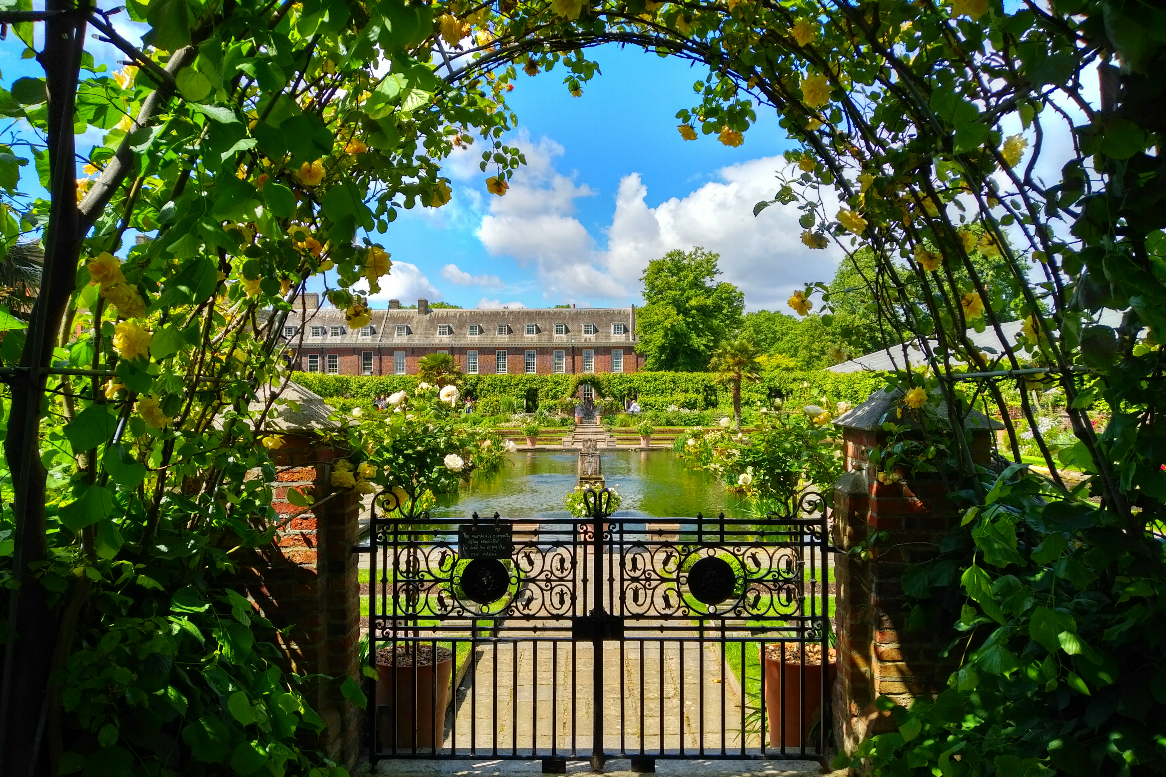 Udsigt gennem blomsterbuet jernport til Kensington Palaces smukke formelle have med dam og historisk rød mursten facade i London