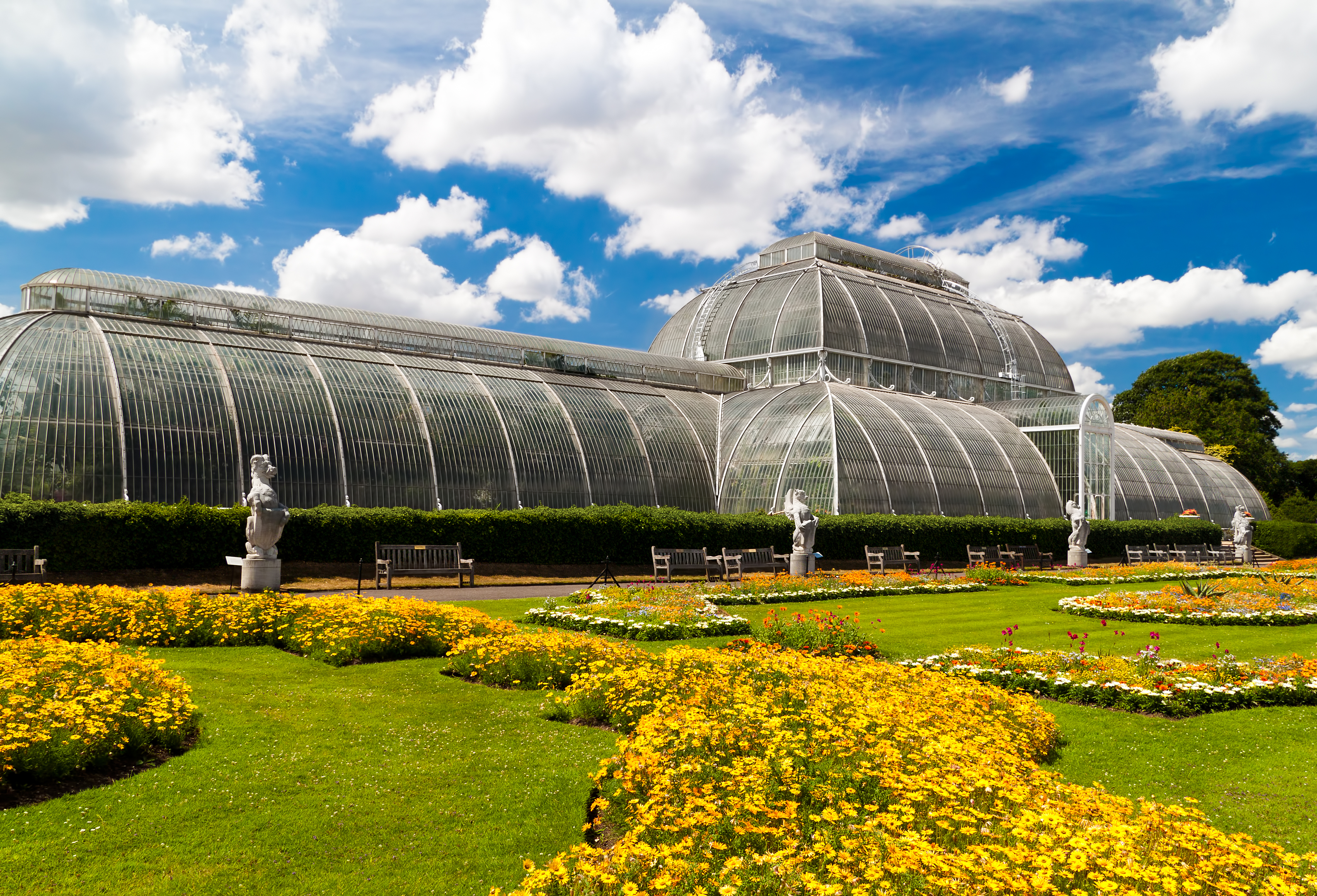 Det historiske victorianske drivhus i Kew Gardens, London, omgivet af velholdte plæner og farverige blomsterbede under en klar blå himmel