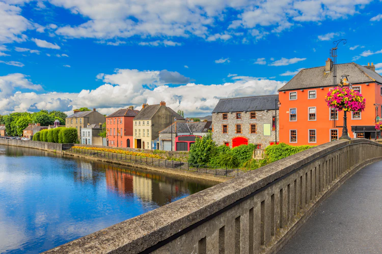 Farverige bygninger langs floden Nore i Kilkenny, Irland, med smukt vejr og blomsterdekorationer på en solrig sommerdag