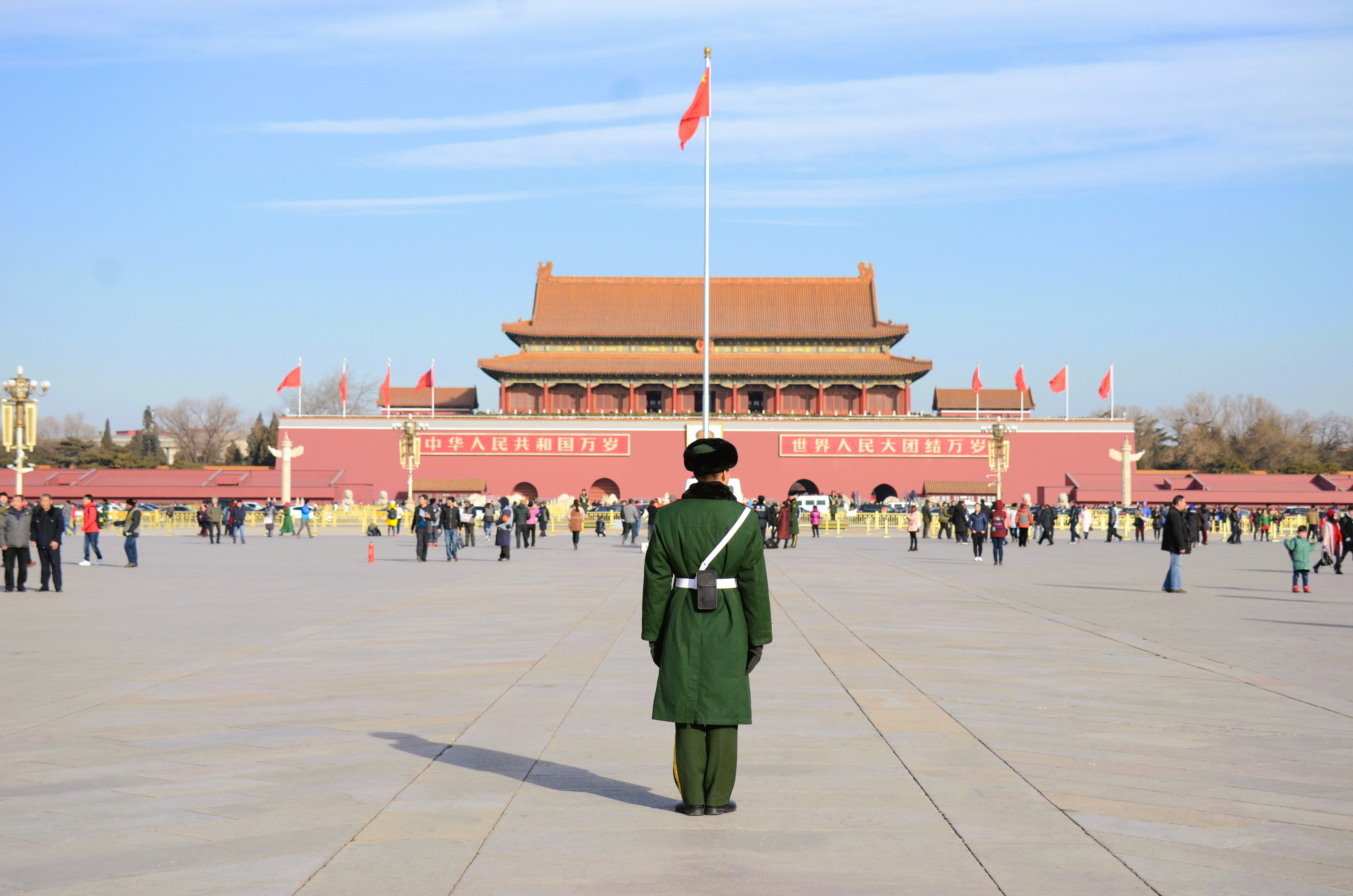 Kinesisk vagt i grøn uniform på Tiananmen Plads i Beijing med Den Forbudte By og rødt flag i baggrunden