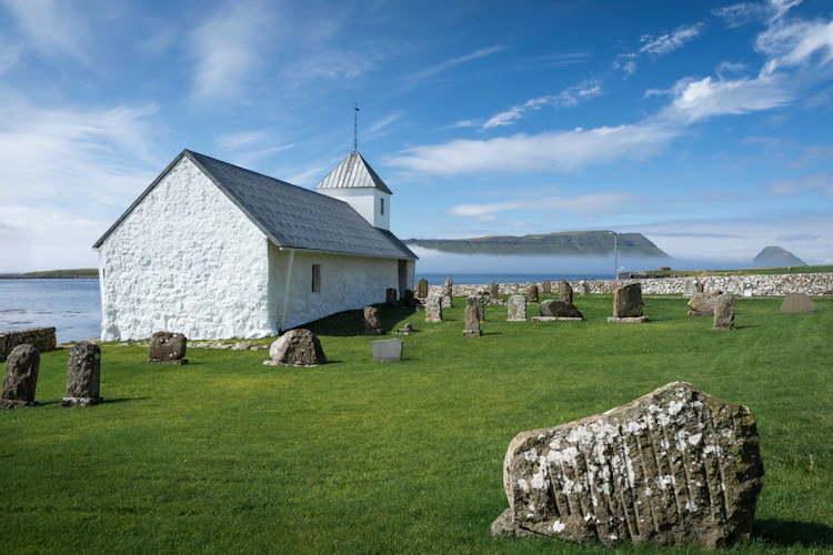 Historisk hvid kirke med gamle gravsten i landsbyen Kirkjubøur på Streymø, Færøerne, med udsigt over Atlanterhavet