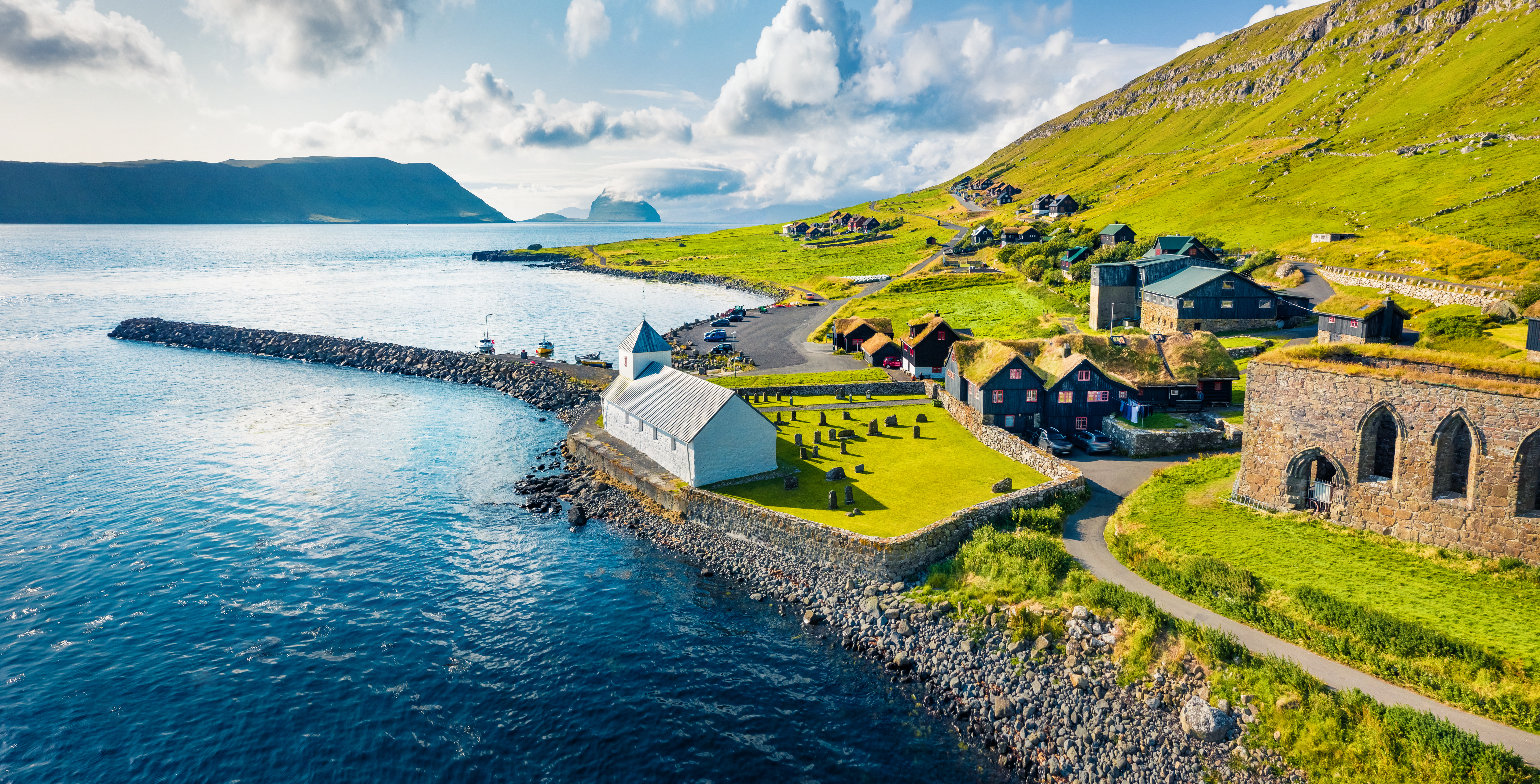 Luftfoto af Kirkjubøur på Færøerne med hvid kirke ved havet, grønne bjerge og Hestur Island i baggrunden