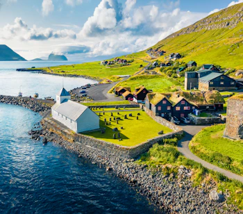 Luftfoto af Kirkjubøur på Færøerne med hvid kirke ved havet, grønne bjerge og Hestur Island i baggrunden