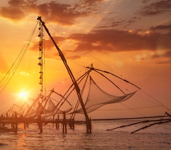Silhuet af traditionelle kinesiske fiskenet mod en dramatisk solnedgang ved Fort Kochi, Kerala, Indien med gyldent lys der oplyser horisonten og skaber en magisk stemning