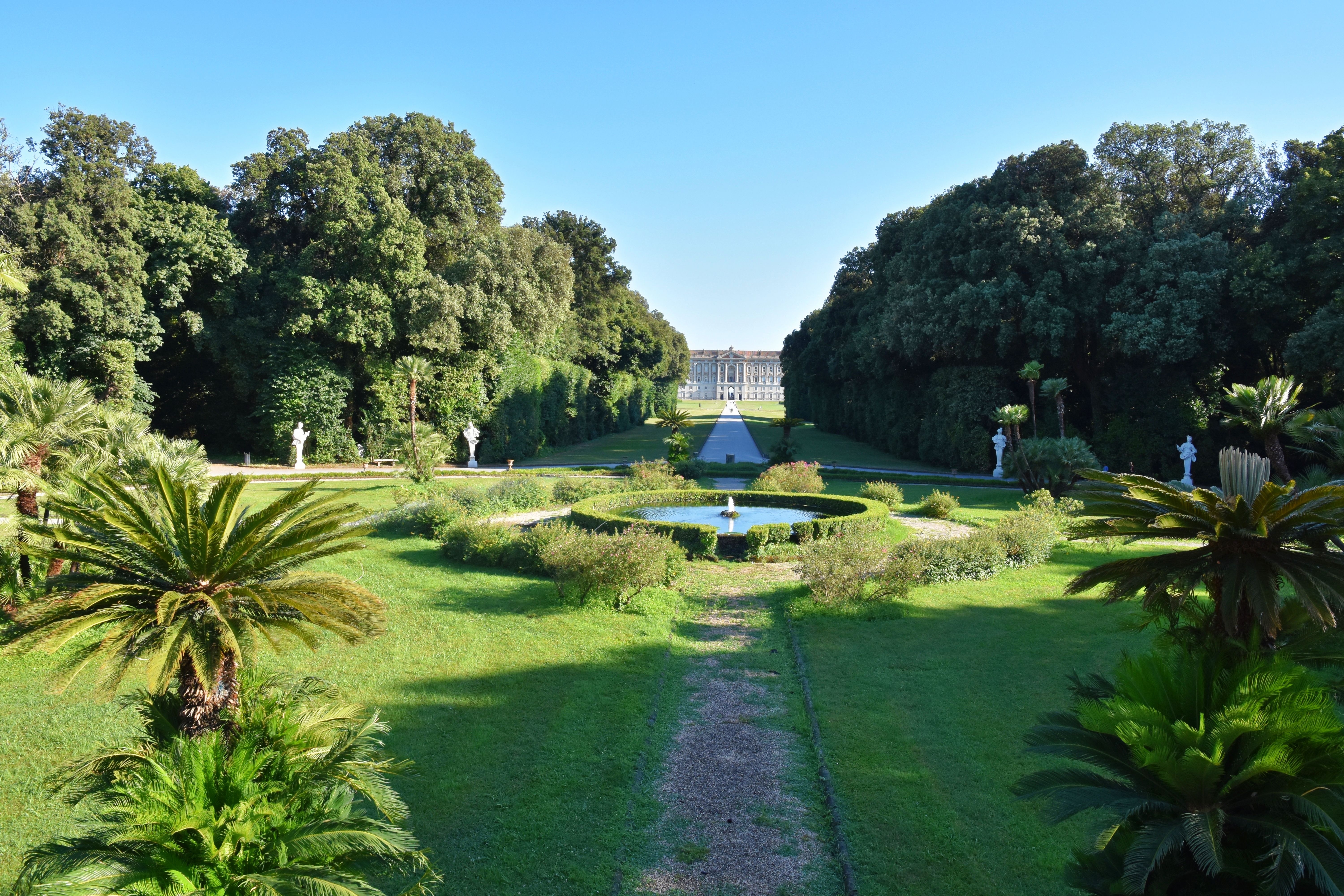 De storslåede barokhaver ved Kongeslottet i Caserta, Italien, med velpleje græsplæner, springvand og palmer der leder blikket mod det majestætiske 1700-tals palads