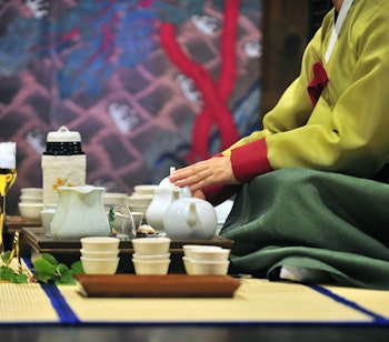 Elegant kvinde i traditionel koreansk hanbok-dragt udfører en teceremoni, hvor hun omhyggeligt hælder te fra en keramisk tepotte i små kopper på et træbord