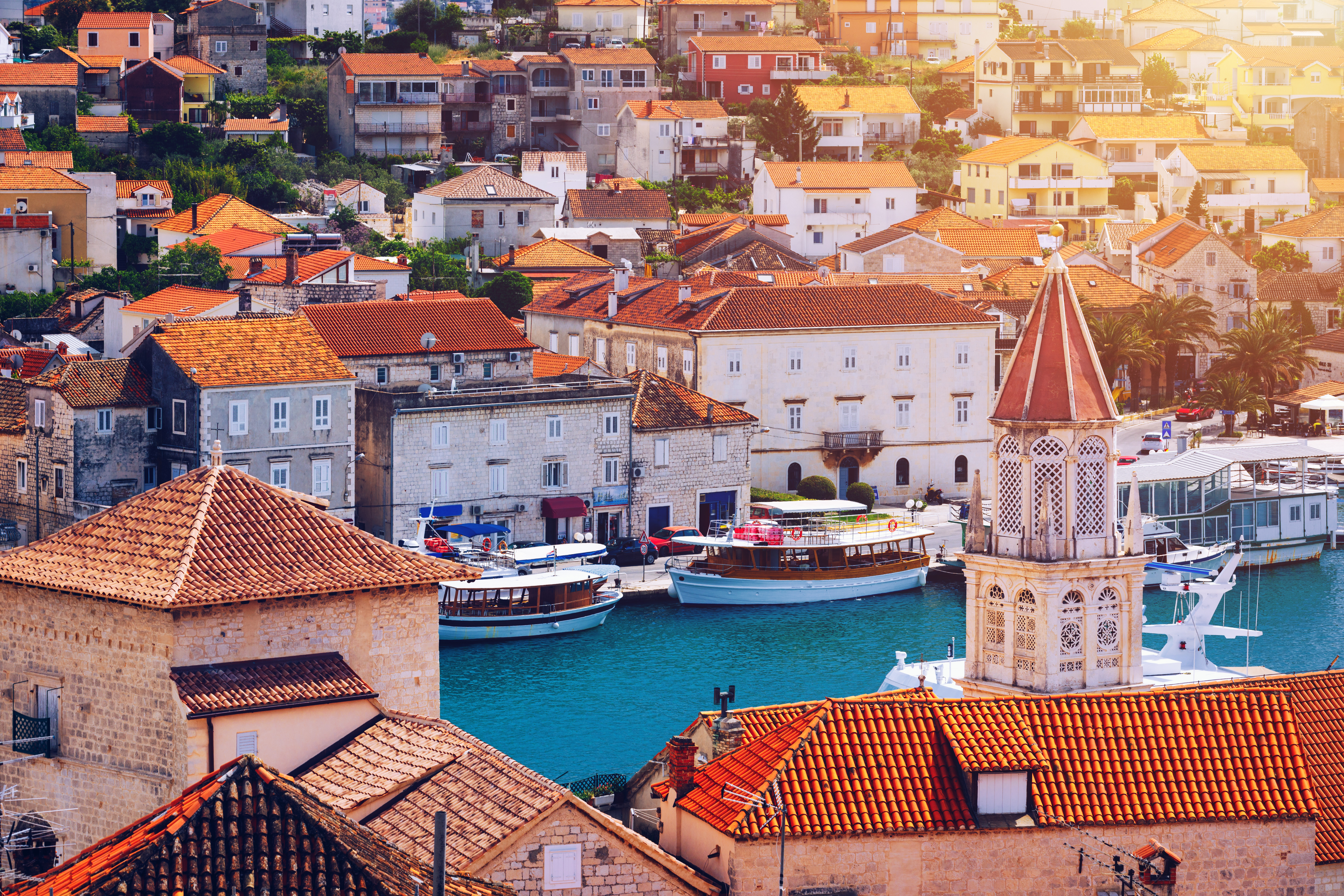 Malerisk kroatisk kystby med røde tegltage, historisk klokketårn og turkisblå havn med traditionelle både ved solnedgang