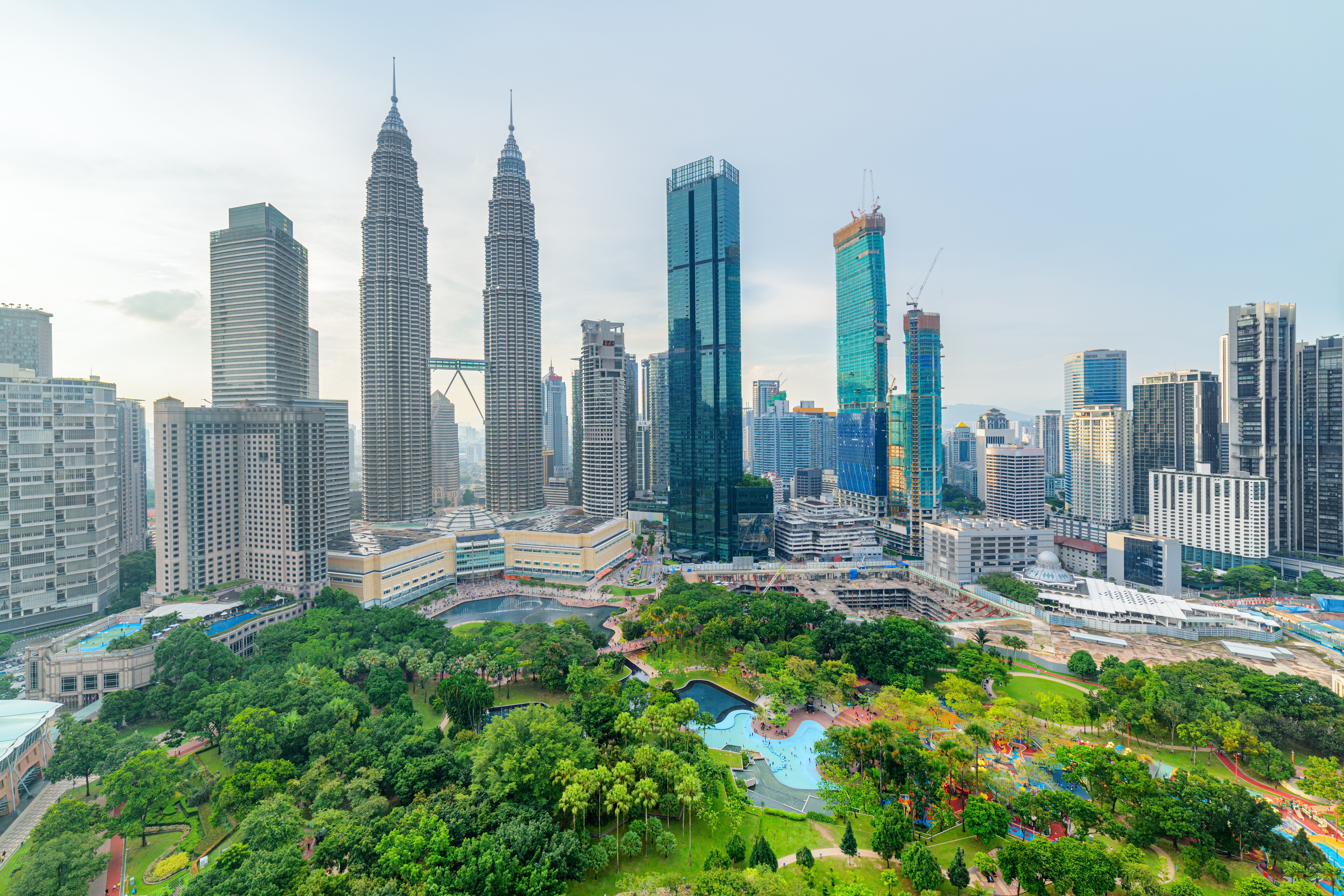 Imponerende luftfoto af Kuala Lumpurs moderne skyline med skyskrabere, der omgiver en frodig grøn bypark under klar blå himmel, Malaysia