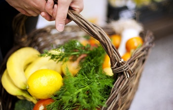 Person bærer flettekurv fyldt med friske frugter og grøntsager, herunder æbler, bananer, citroner og urter på lokalt marked