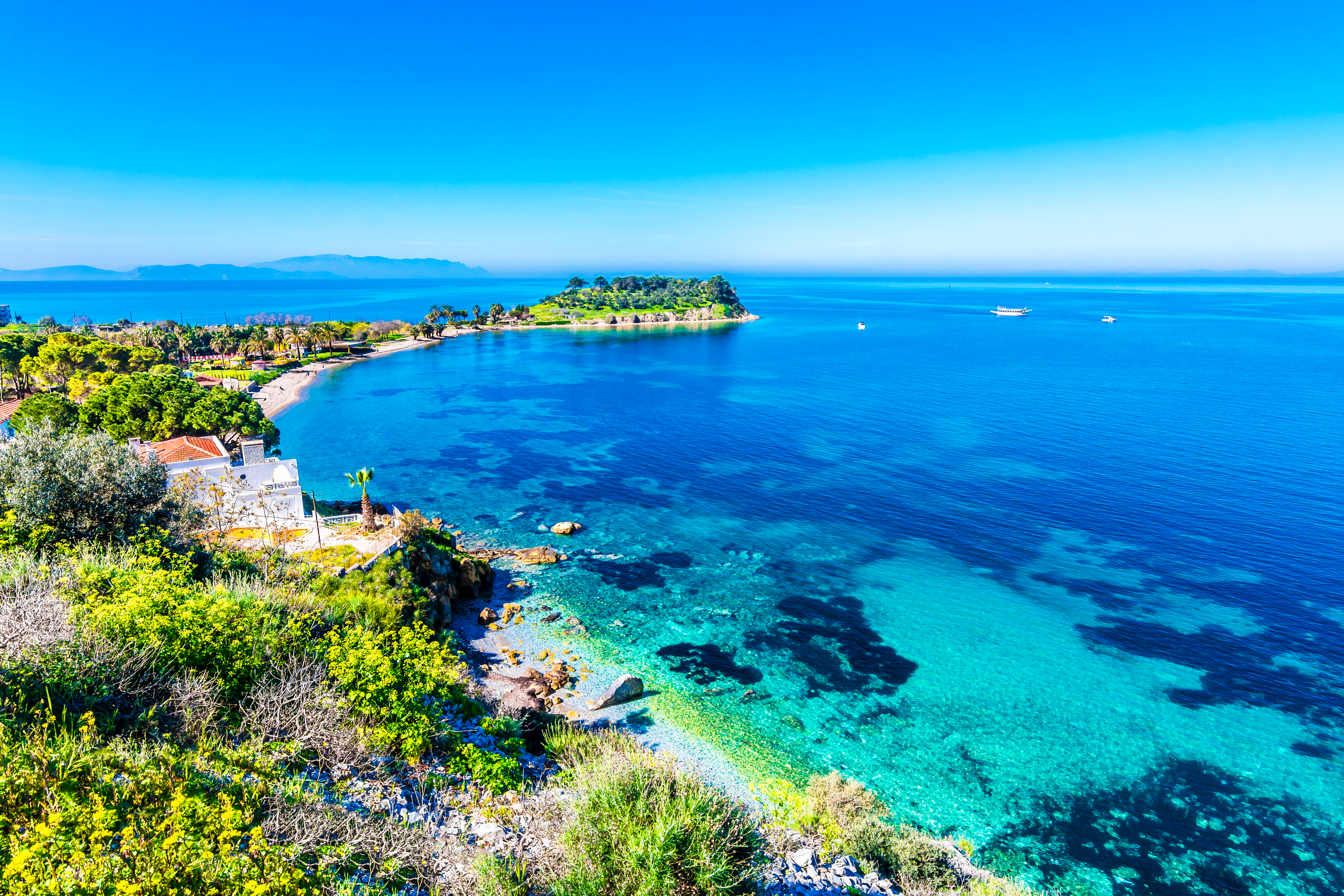 Betagende udsigt over Kusadasis kystlinje i Tyrkiet, med det turkisblå Ægæiske Hav, den historiske halvø og den maleriske havn omgivet af grønne bakker under en klar blå himmel