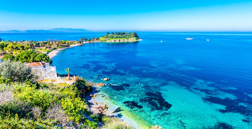 Betagende udsigt over Kusadasis kystlinje i Tyrkiet, med det turkisblå Ægæiske Hav, den historiske halvø og den maleriske havn omgivet af grønne bakker under en klar blå himmel