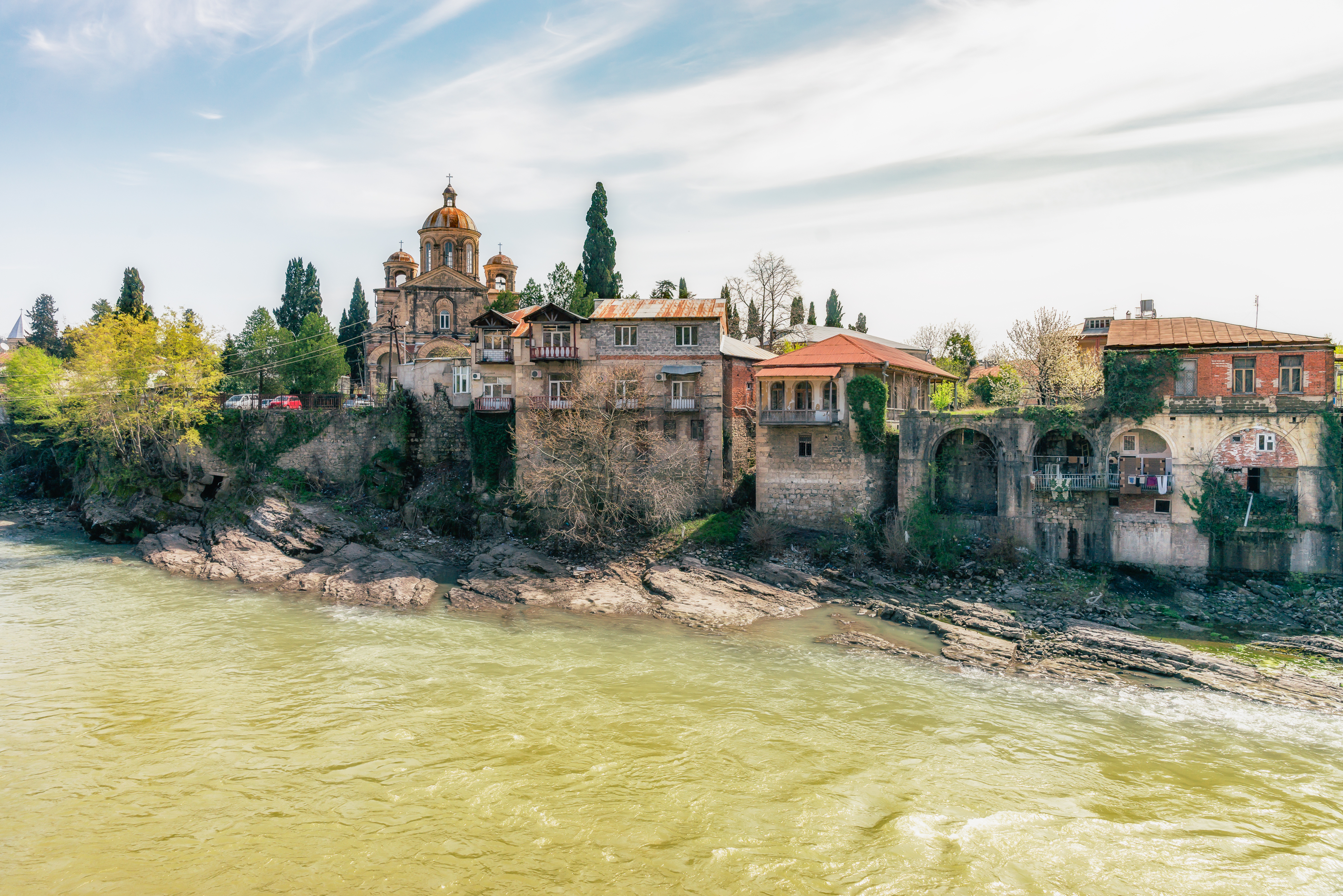 Malerisk udsigt over Rioni-floden der snor sig gennem den historiske by Kutaisi i Georgien med traditionelle stenbygninger og de grønne Kaukasus-bjerge i baggrunden