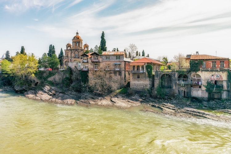 Malerisk udsigt over Rioni-floden der snor sig gennem den historiske by Kutaisi i Georgien med traditionelle stenbygninger og de grønne Kaukasus-bjerge i baggrunden