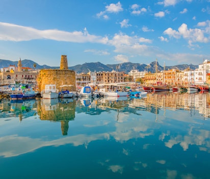 Idyllisk udsigt over Kyrenias gamle havn i Nordcypern med farverige både, historiske bygninger og den middelalderlige fæstning spejlet i det rolige Middelhavs azurblå vand