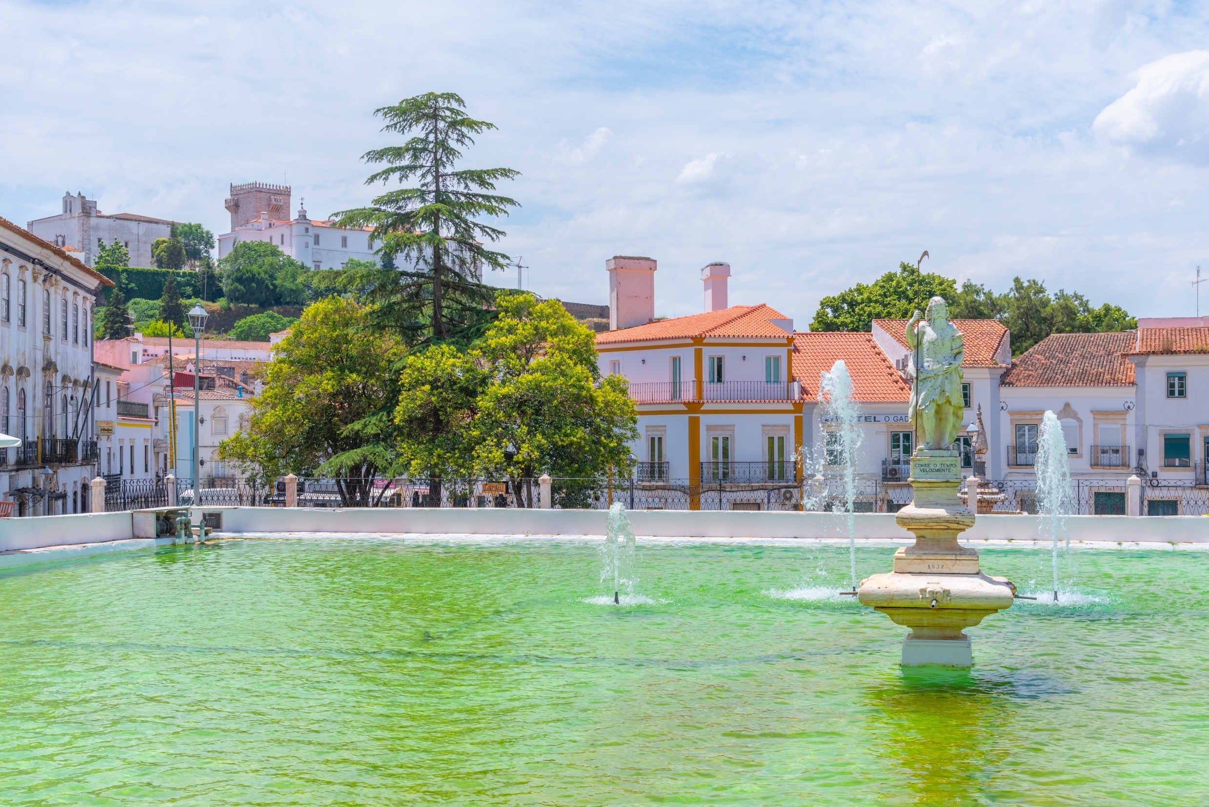 Det smukke Lago da Gadanha i Estremoz, Portugal, med grønt vand, en skulpturel fontæne og traditionelle portugisiske bygninger med orange tagtegl i baggrunden