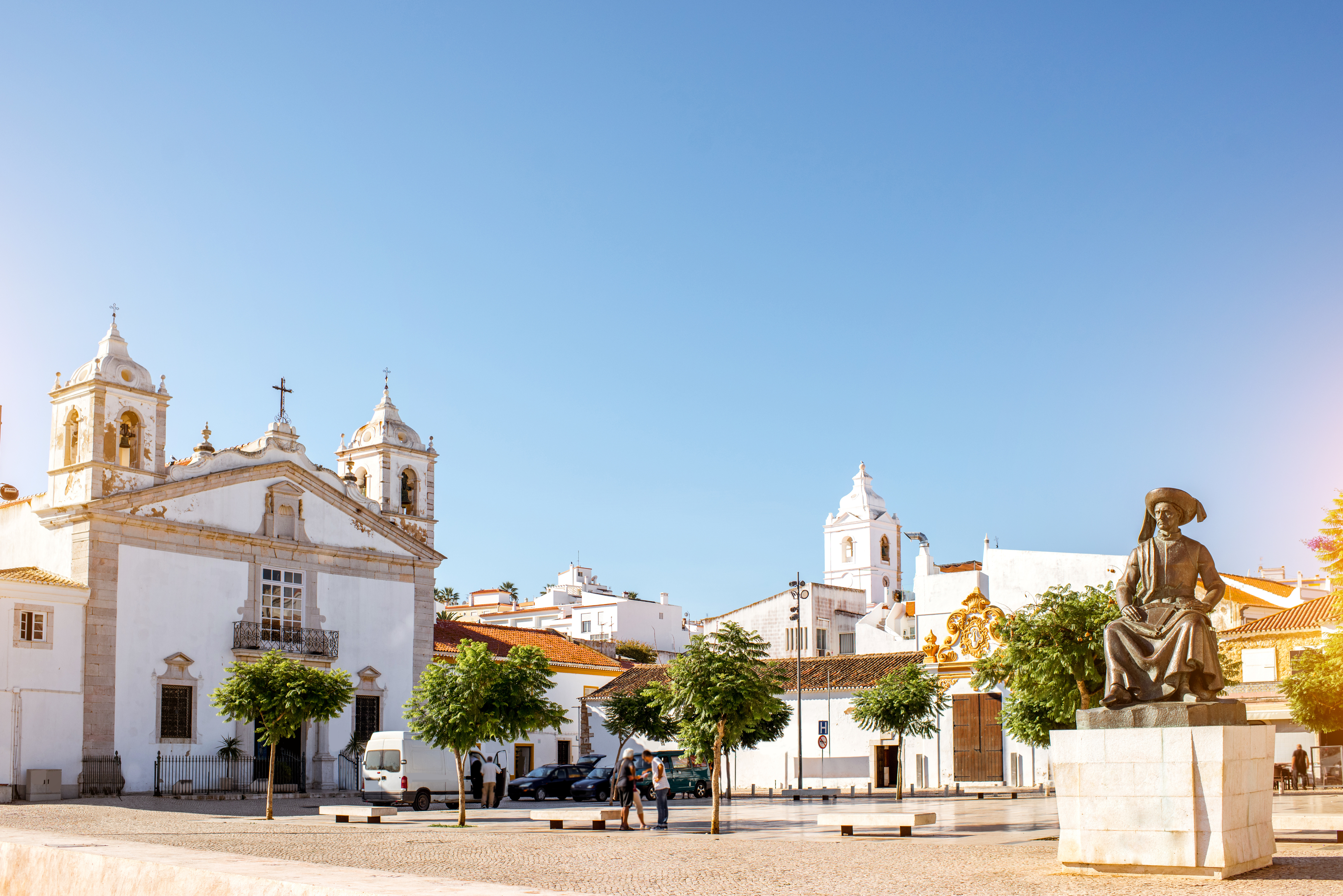 Solrig plads i Lagos, Portugal, med den hvide Santa Maria Kirke, en bronzestatue og charmerende Algarve-arkitektur under en klar blå himmel