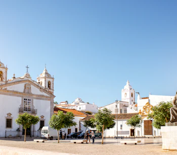 Solrig plads i Lagos, Portugal, med den hvide Santa Maria Kirke, en bronzestatue og charmerende Algarve-arkitektur under en klar blå himmel