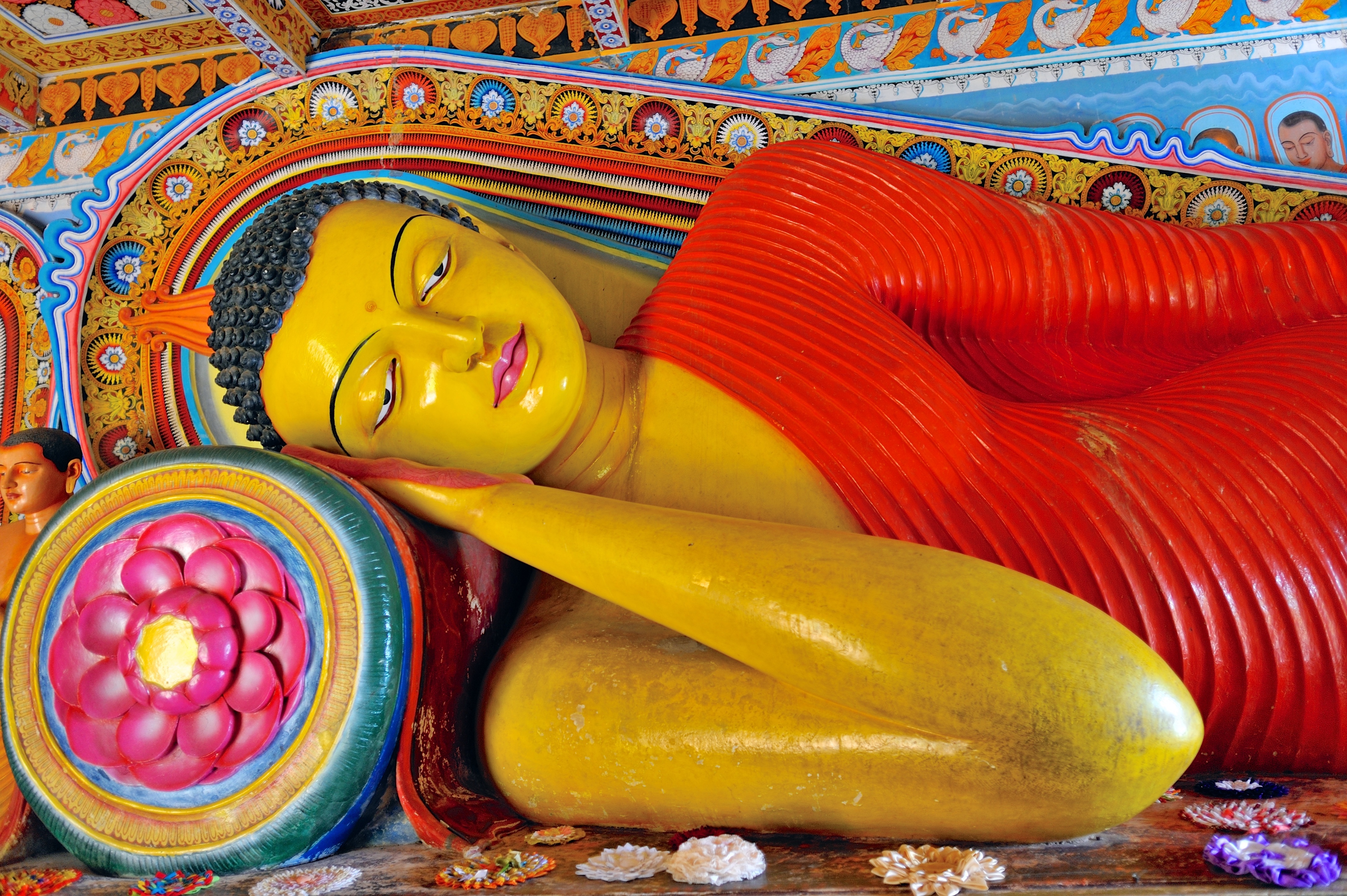 Farverig liggende Buddha-statue med rød klædning i Isurumuniya-templet, Anuradhapura, Sri Lanka, omgivet af traditionelle dekorative mønstre