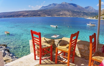 Traditionel græsk taverna med røde stole ved det krystalklare turkisblå hav i den charmerende landsby Limeni på Peloponnes - en idyllisk frokostplads med udsigt over bugten og bjergene