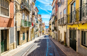 Charmerende smal gade i Lissabons historiske Alfama-kvarter med traditionelle bygninger, sporvognsskinner og en gul sporvogn der kører op ad den stejle bakke på en smuk solrig sommerdag