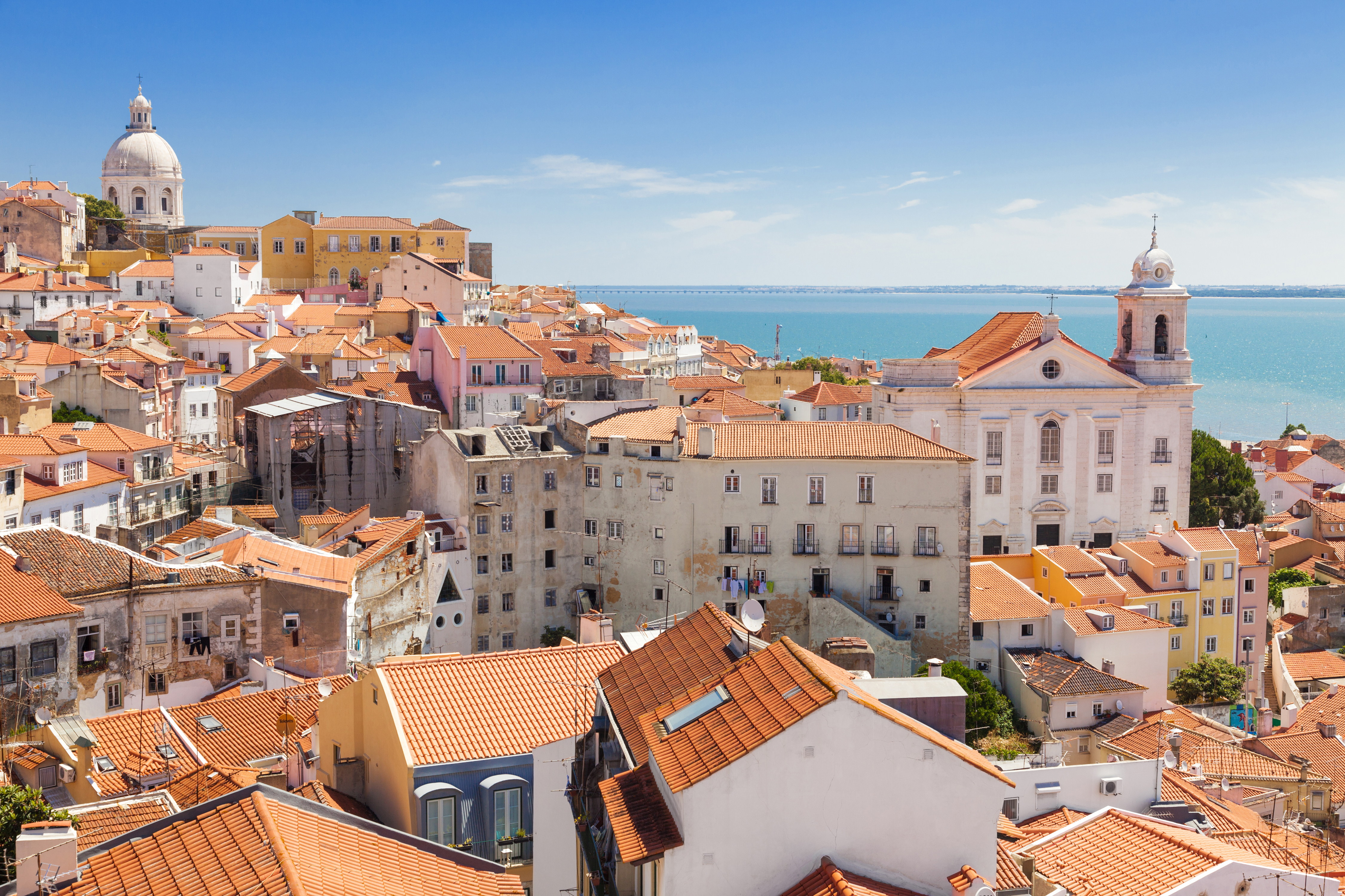 Panoramaudsigt over Alfama-kvarteret i Lissabon med karakteristiske røde tagsten, hvid kirke og Tejo-floden i baggrunden