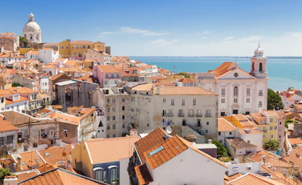 Panoramaudsigt over Alfama-kvarteret i Lissabon med karakteristiske røde tagsten, hvid kirke og Tejo-floden i baggrunden