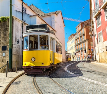 Charmerende gul vintage sporvogn der kører gennem de smalle brostensbelagte gader i Lissabons historiske centrum, omgivet af farverige traditionelle bygninger på en solrig sommerdag