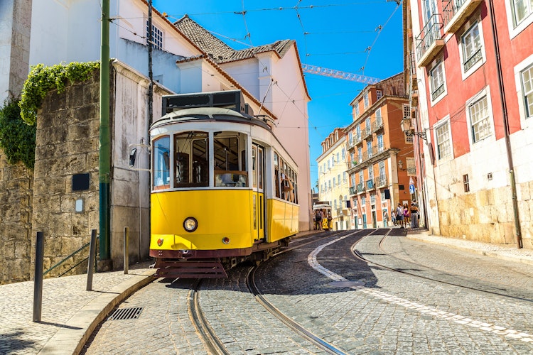 Charmerende gul vintage sporvogn der kører gennem de smalle brostensbelagte gader i Lissabons historiske centrum, omgivet af farverige traditionelle bygninger på en solrig sommerdag