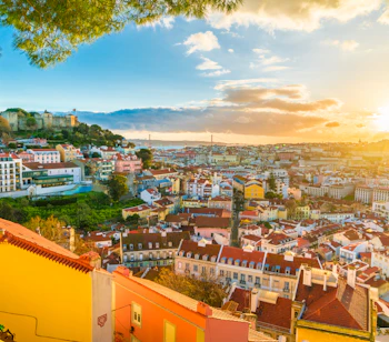 Betagende panoramaudsigt over Lissabons historiske bylandskab med Alfama-kvarteret og São Jorge-slottet badet i solnedgangens gyldne lys