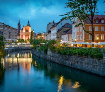 Den historiske Tromostovje-bro over Ljubljanica-floden i Ljubljanas charmerende centrum ved aftentid, med smukke bygninger og vandspejlinger