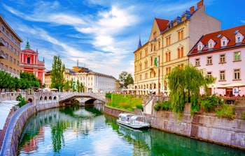 Den ikoniske Tromostovje-bro oplyst om aftenen med den franciskanske kirke spejlet i Ljubljanica-floden i Ljubljanas historiske centrum