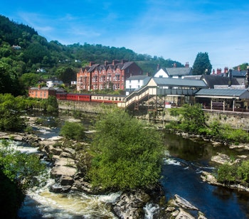 Den historiske Llangollen jernbanestation med den smukke flod Dee i forgrunden og traditionel walisisk arkitektur under en klar blå himmel