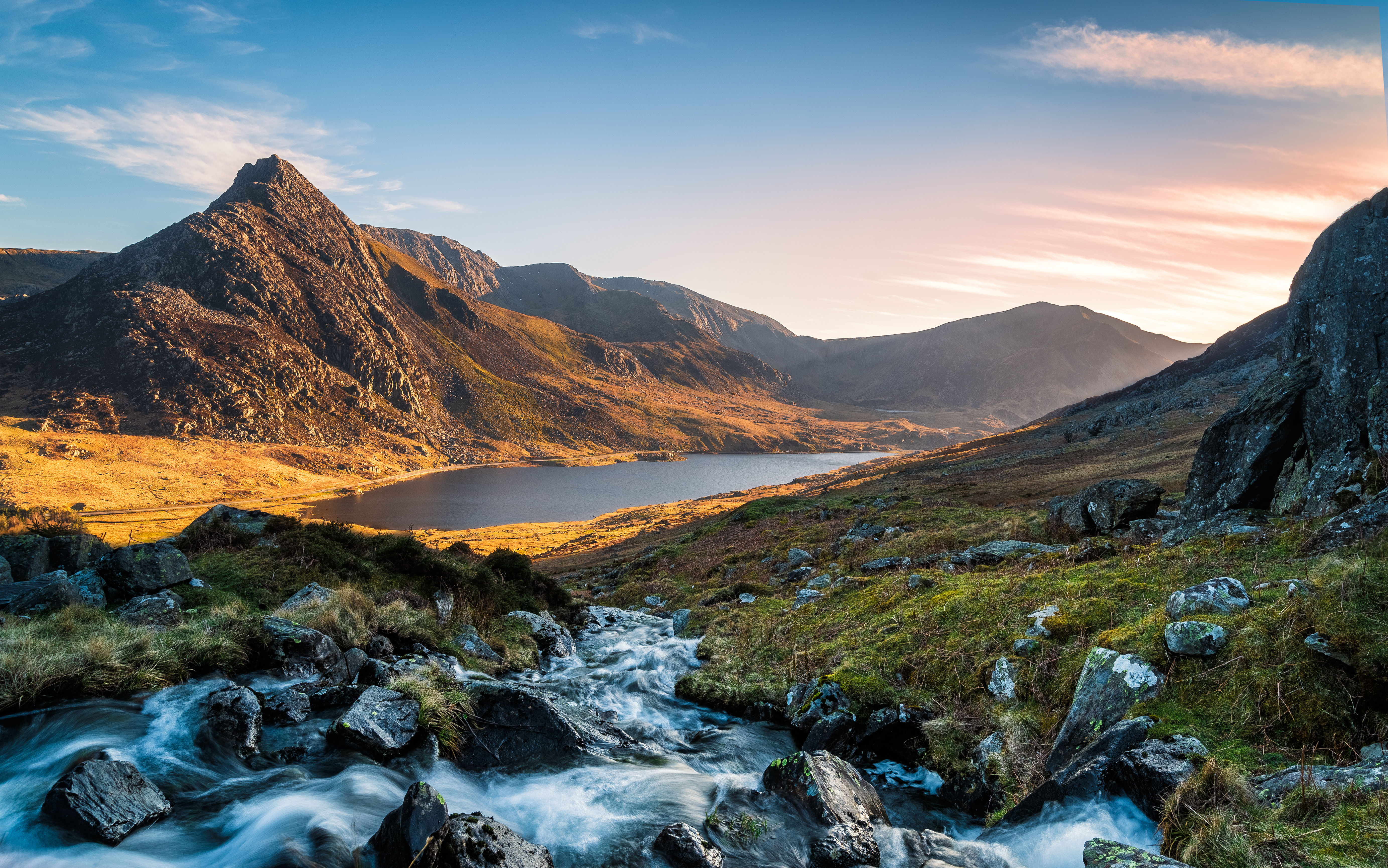 Betagende solnedgang over Llyn Ogwen søen i Snowdonia Nationalpark, Wales, med dramatiske bjergtoppe og gylden himmel der spejler sig i det rolige vand