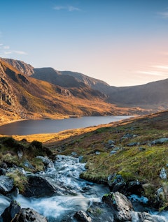 Betagende solnedgang over Llyn Ogwen søen i Snowdonia Nationalpark, Wales, med dramatiske bjergtoppe og gylden himmel der spejler sig i det rolige vand