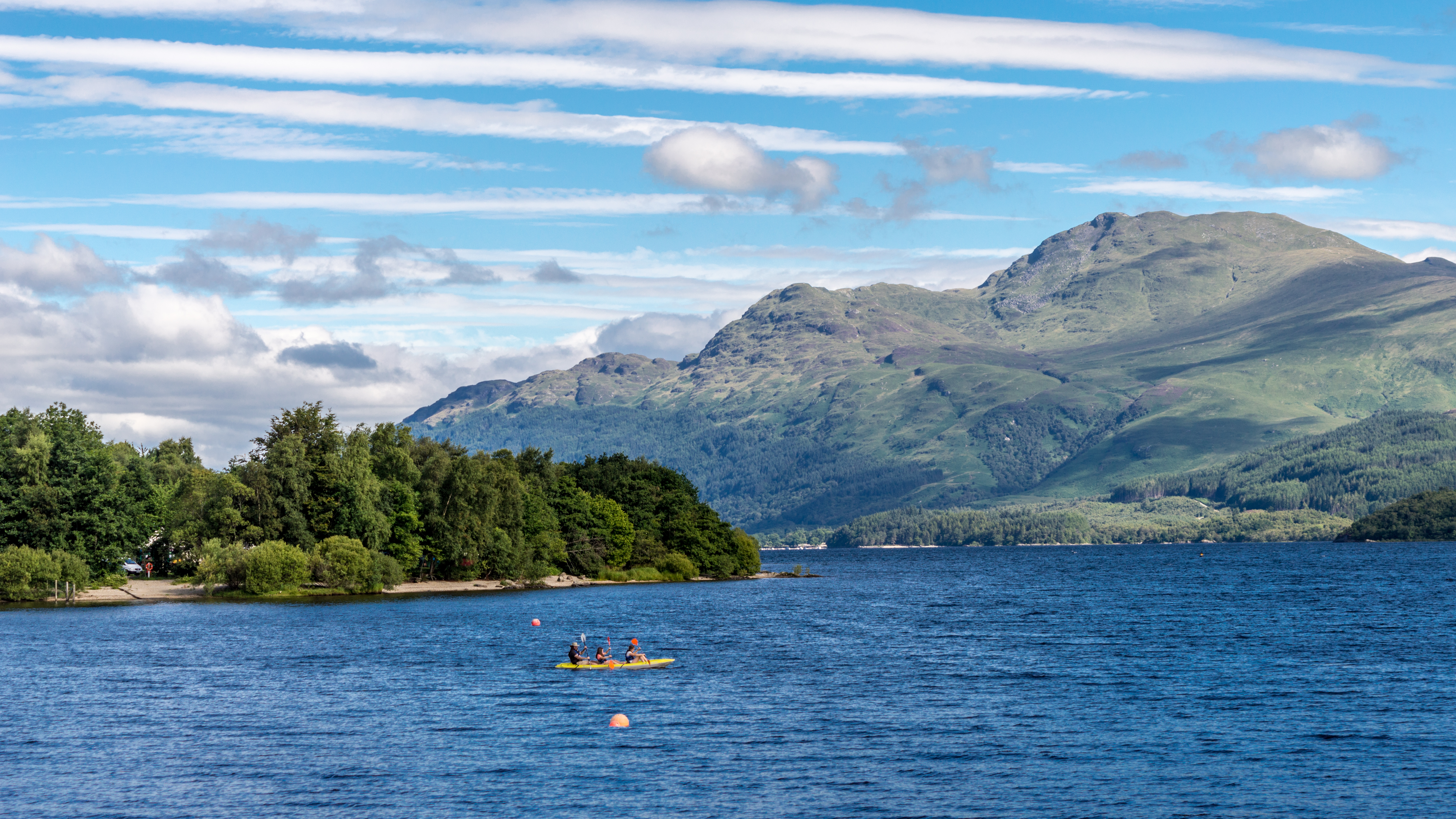 Turister i kajak på Loch Lomond med skotske højlande og grønne skove i baggrunden på solrig sommerdag