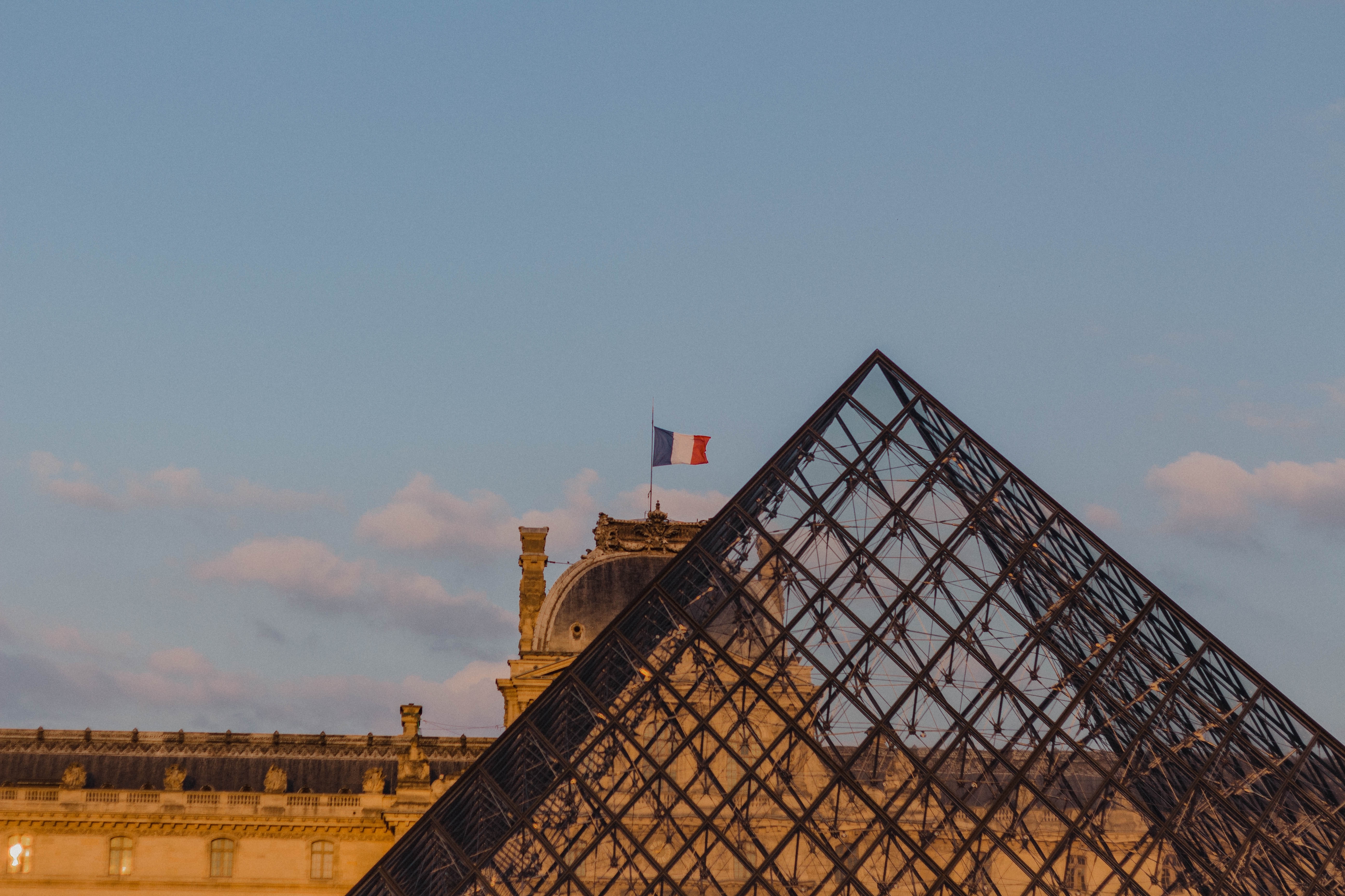 Den ikoniske glaspyramide ved Louvre-museet i Paris med det historiske palads og det franske flag mod en blå himmel